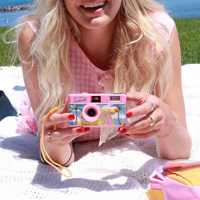 A woman holding a Malibu Barbie 35mm film camera while laying on a blanket in the grass outside. 