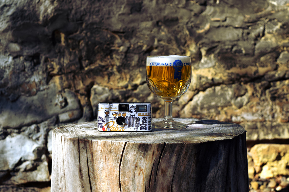 The Pabst Blue Ribbon film camera next to a goblet of beer on a tree stump in the woods. 